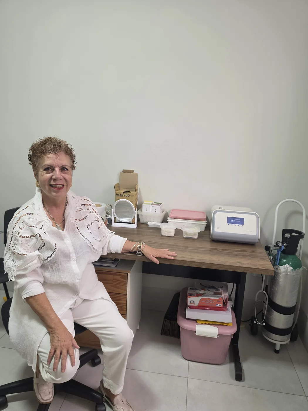 Beth Perez sentada em seu consultório de ozonioterapia, ao lado do gerador de ozônio e cilindro de oxigênio
